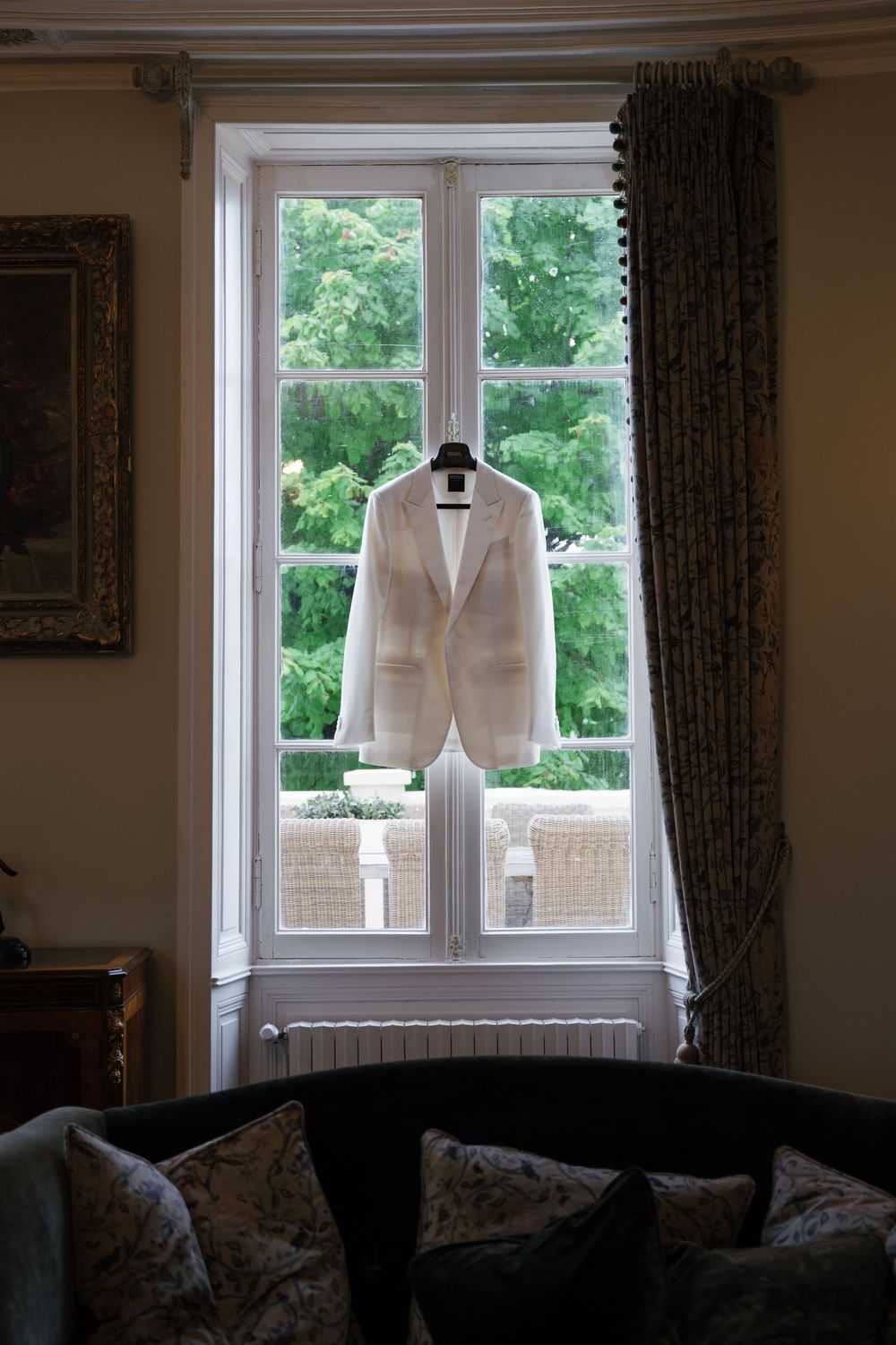 A white suit jacket hangs by a window in an elegant room with dark curtains.