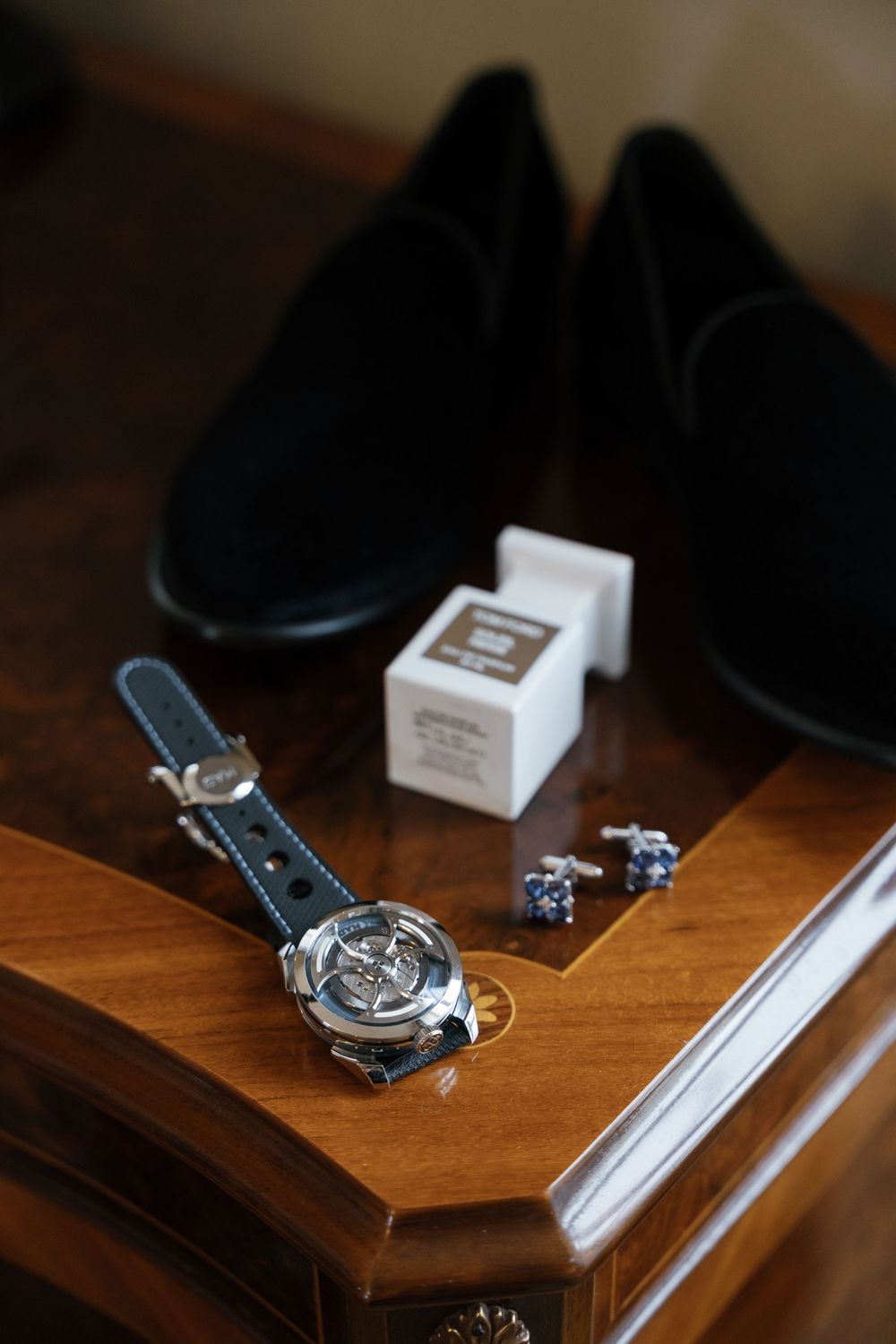 Groom's accessories including black dress shoes, watch, cufflinks and ring box arranged on wooden surface.