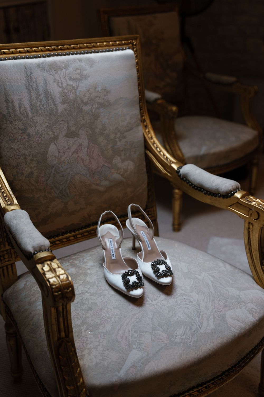 Silver high heeled shoes rest on an ornate gold and white antique chair.