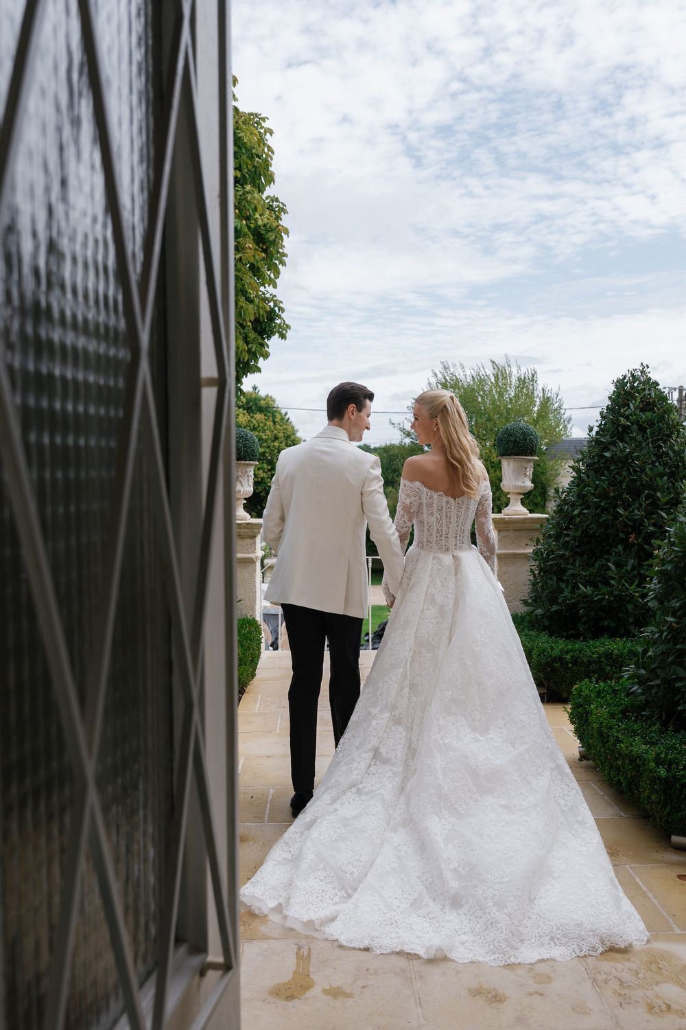 A couple in wedding attire walks together on a stone terrace with lush greenery and cloudy skies in the background.