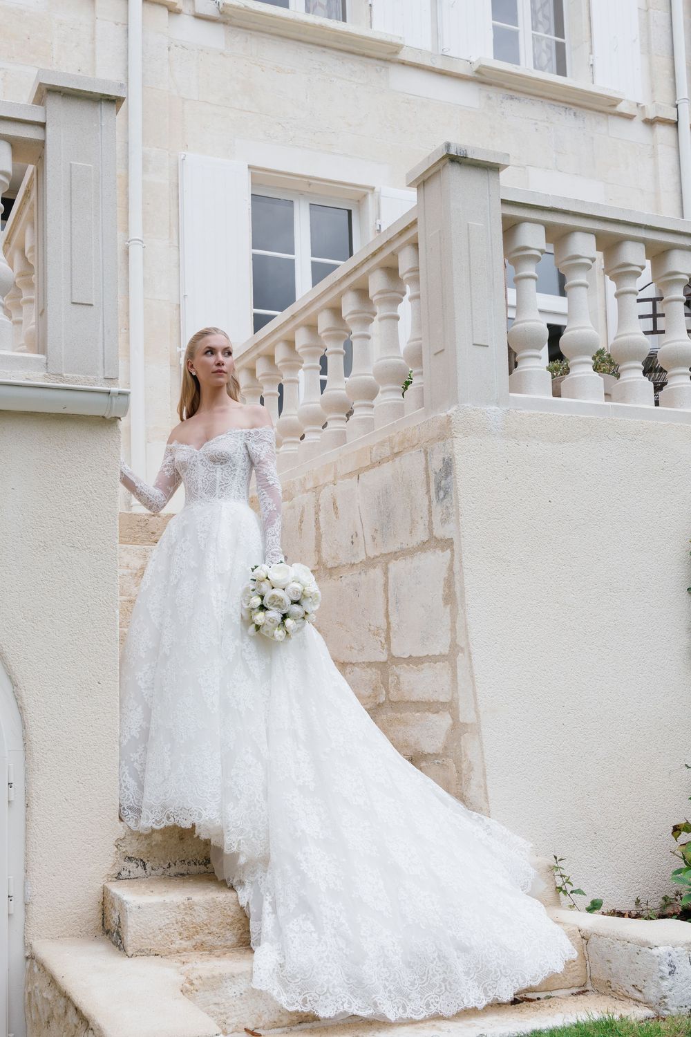 Bride in flowing lace wedding gown poses gracefully on stone staircase of historic building.