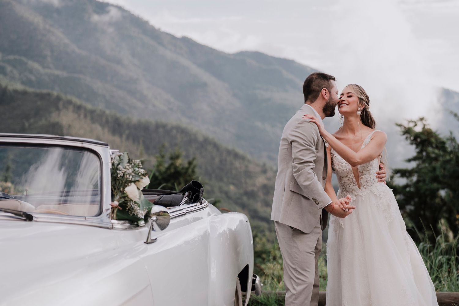 Couple shares intimate moment near vintage car with mountains in background.