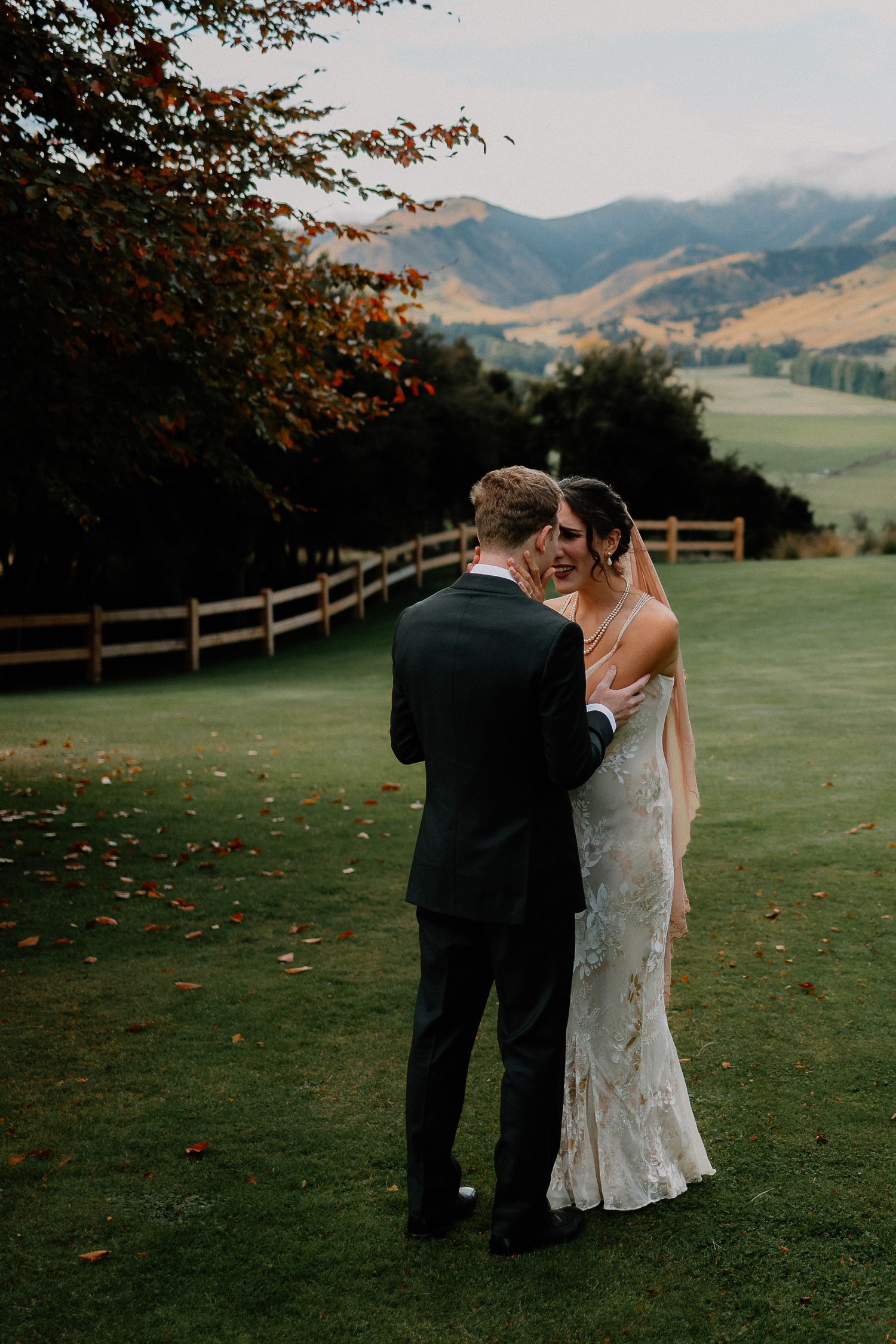 A couple in formal wedding attire share an intimate moment on a scenic hillside with mountain views and wooden fencing.