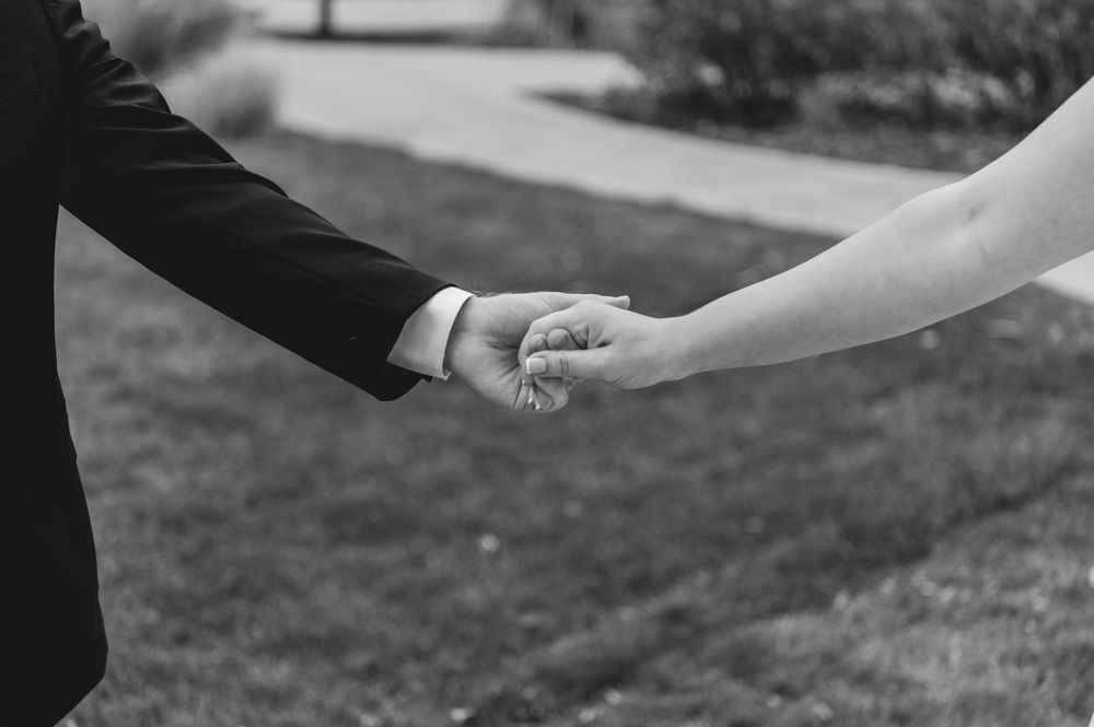 A couple holding hands in formal wedding attire walk together outdoors in a black and white photograph.