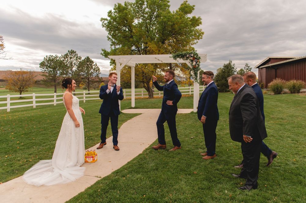 Wedding party gathered on a grassy lawn with white fencing and fall foliage in the background.