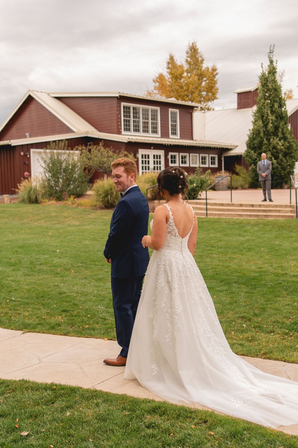 A couple in wedding attire walk together on a grassy hill outside a rustic brown building on their special day.