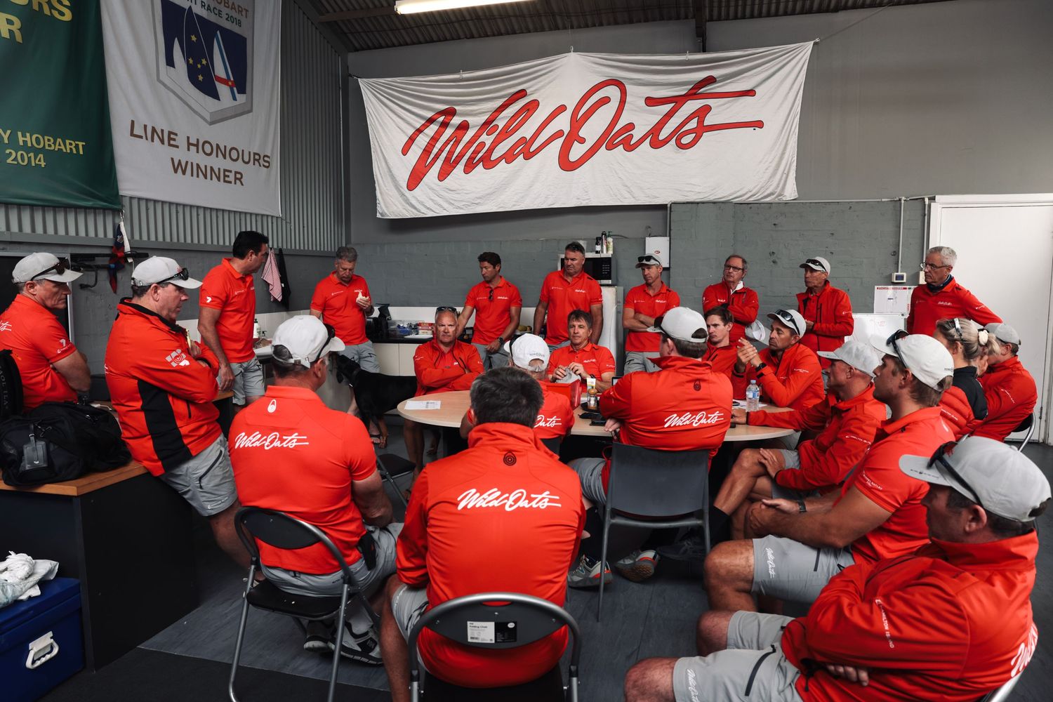 Racing team crew members in red uniforms gathered for meeting in workshop.