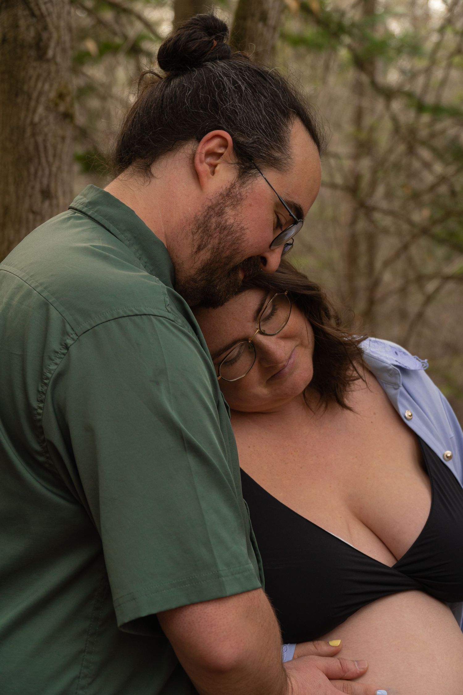 A bearded man in a green shirt tenderly embraces a pregnant woman outdoors among trees.