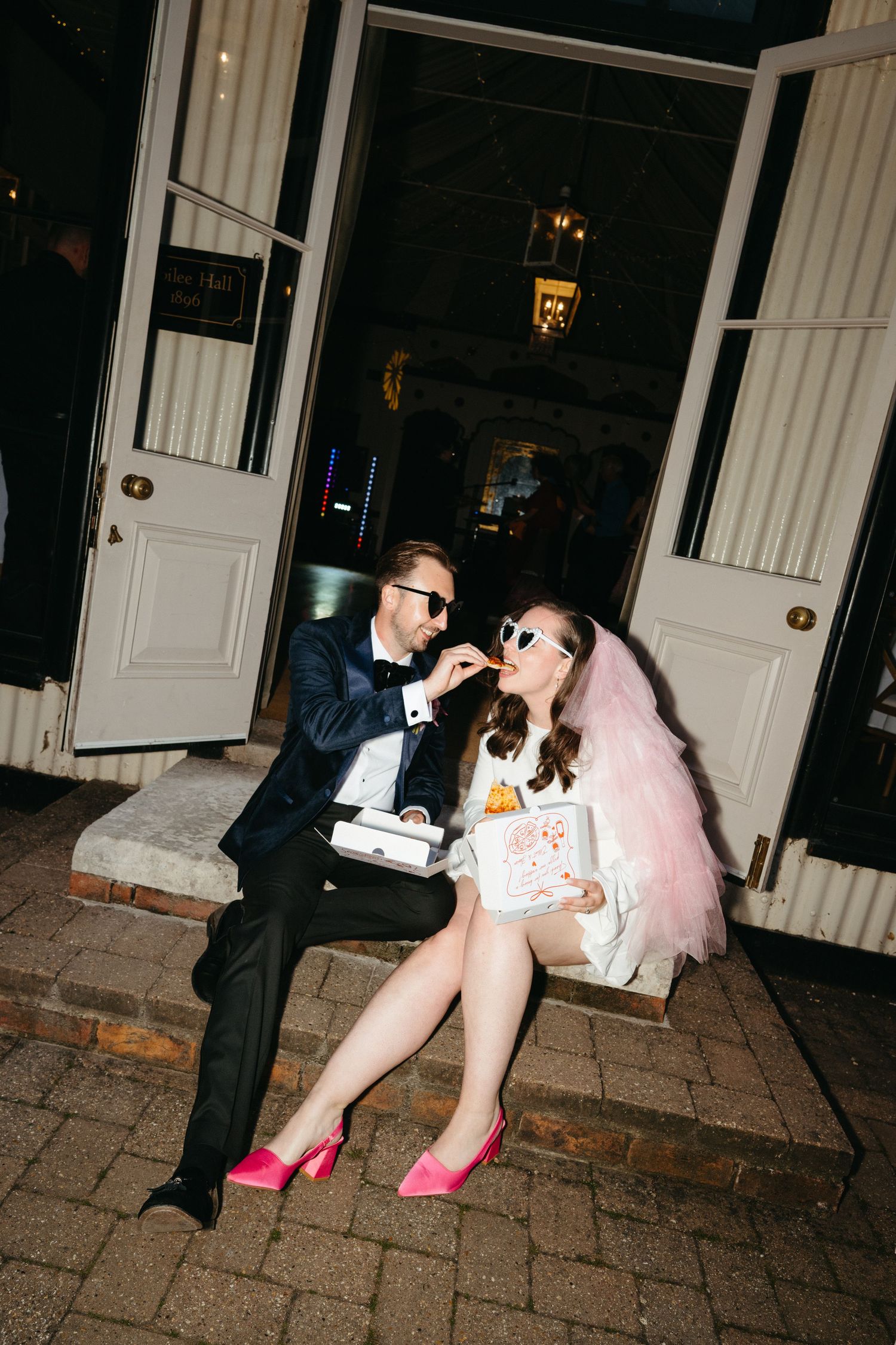 A couple in formal attire playfully posing on brick pavement outside a building with white columns.Taken by editorial wedding photographer.