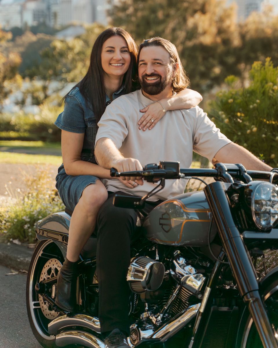 A couple sits together on a Harley-Davidson motorcycle during a golden hour photoshoot in a park setting.