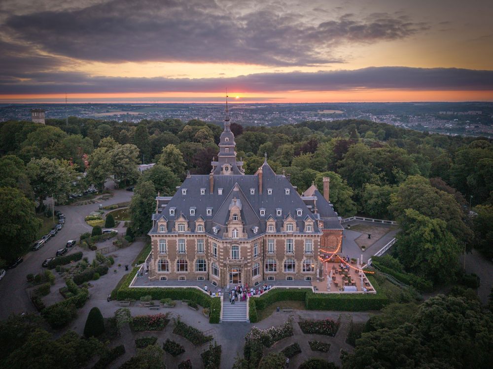 Aerial view of an ornate French chateau-style mansion with manicured gardens at sunset against a colorful sky.