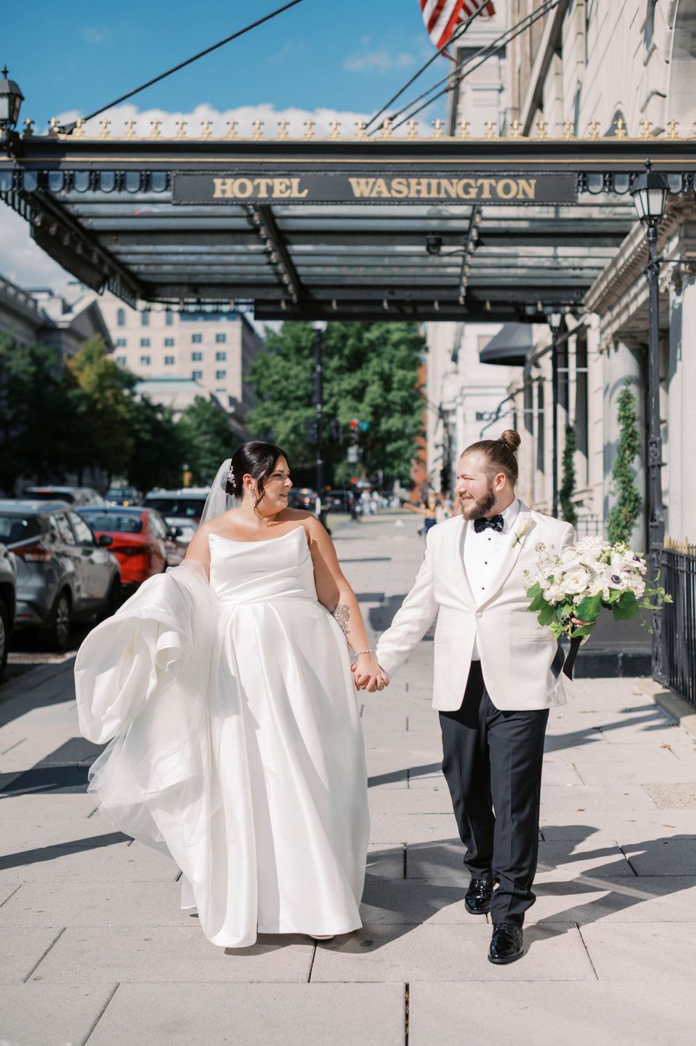 A bride and groom in white attire dance and twirl together on a sidewalk outside the Hotel Washington.