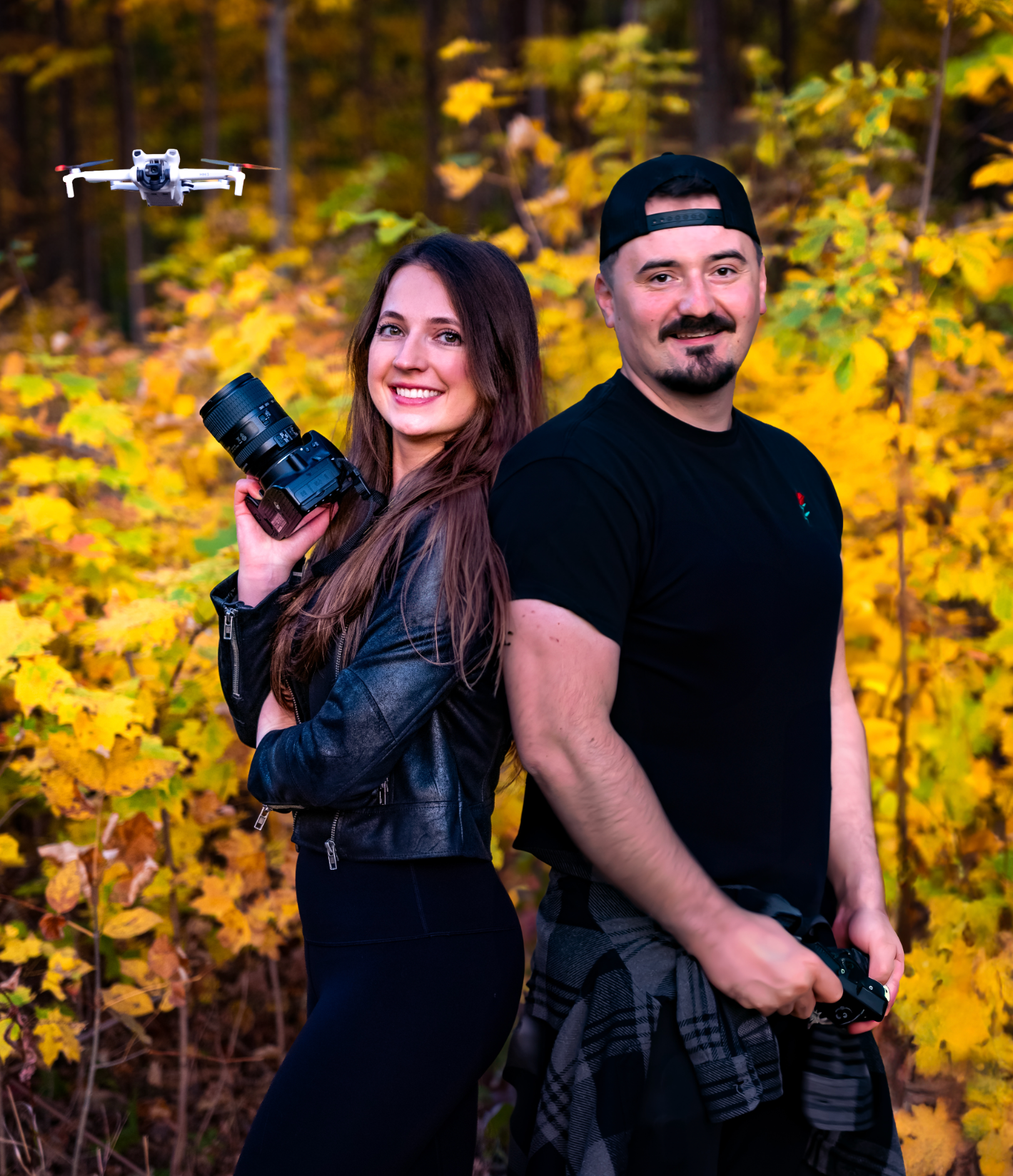 A couple poses together in autumn foliage with cameras during an outdoor photography session.