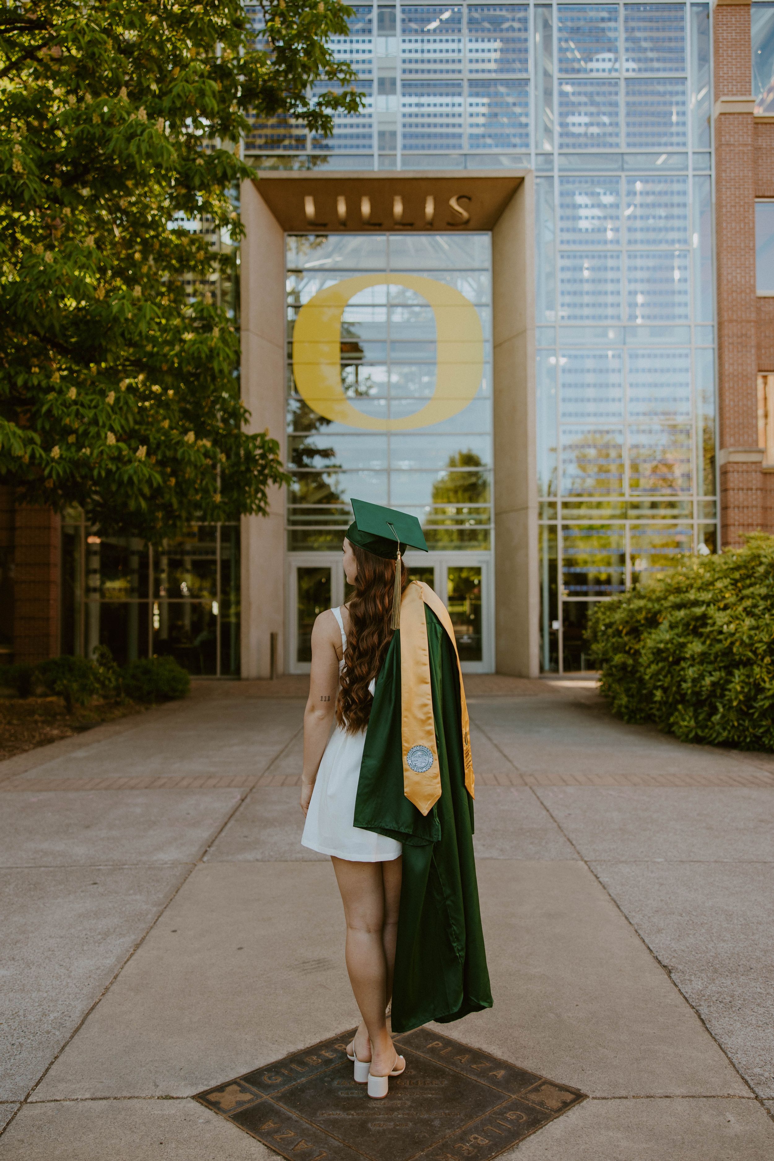 Graduate in green cap and gown standing in front of University of Oregon entrance.