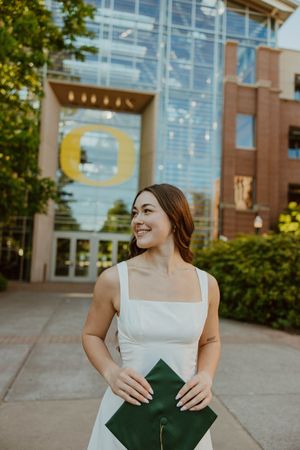 Graduate holding green diploma cover stands outside University of Oregon building.