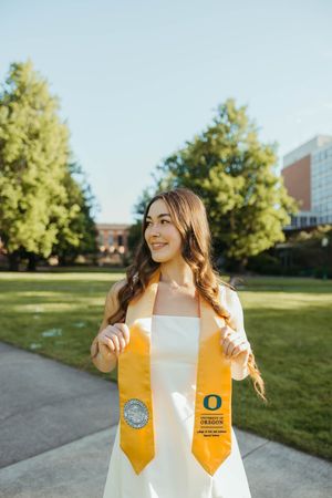 Graduate displaying yellow graduation stole with Oregon logo on campus lawn.