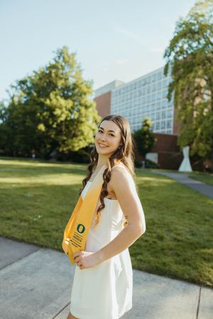 Graduate in white dress with yellow stole poses on University of Oregon campus.