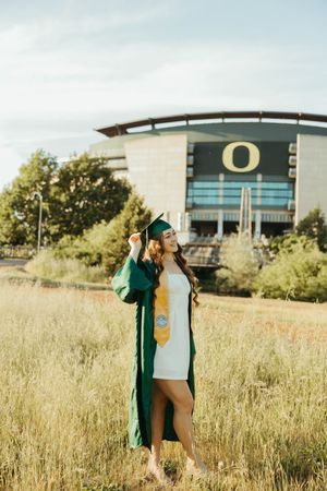 Graduate in green cap and gown celebrates in front of University of Oregon stadium on sunny day.