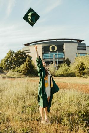 Graduate tosses green cap in celebration with University of Oregon building in background.