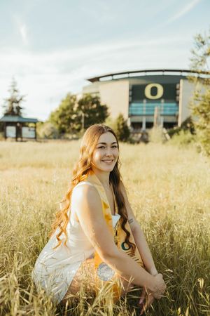 Graduate sits in golden field with university building visible in distance.