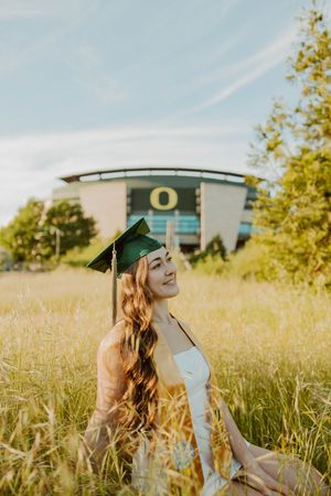 Graduate in cap poses joyfully in sunny field with campus building behind.