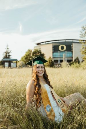 Graduate in green regalia smiles while sitting in sunlit field near university.