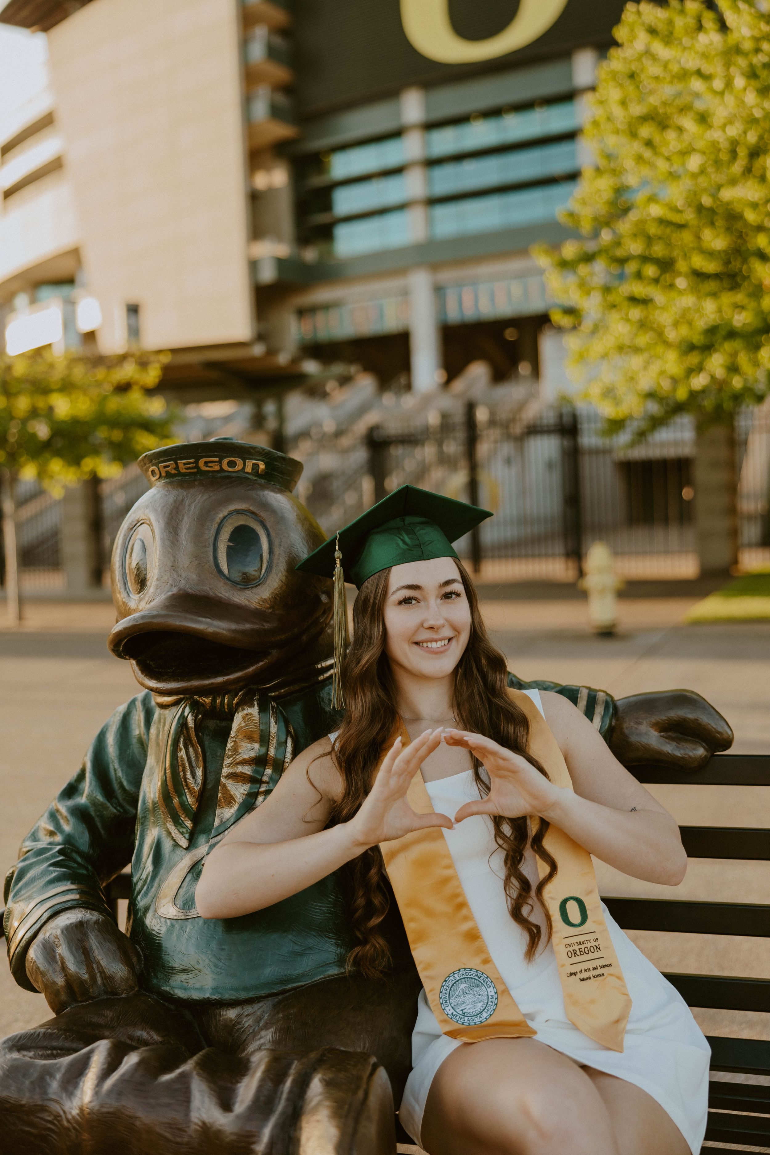 Graduate poses with Oregon Ducks mascot statue on campus bench.