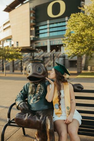 Graduate sharing moment with Oregon Duck mascot statue outside stadium.