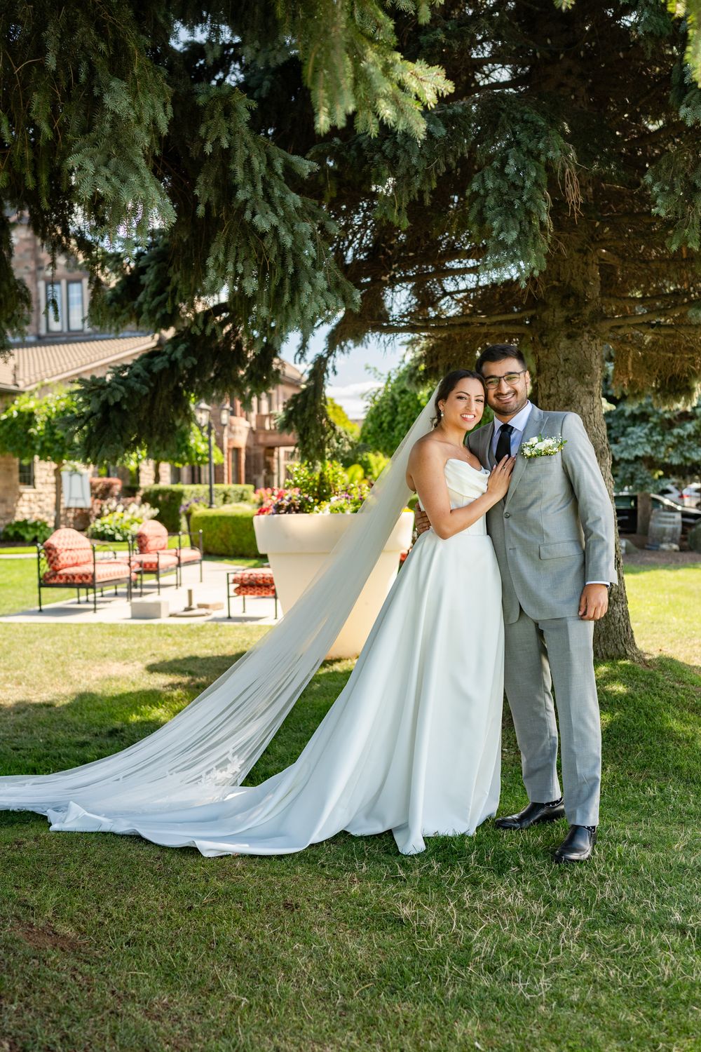A wedding couple poses on a grassy lawn with evergreen trees in the background.