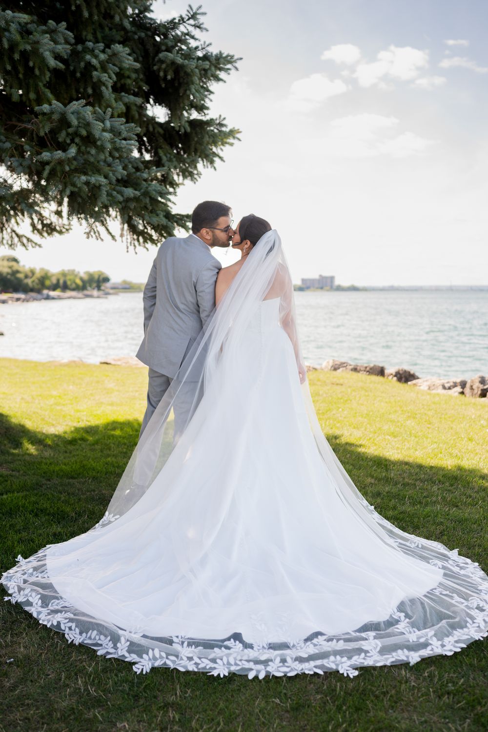 A wedding dress with intricate lace train spreads beautifully across green grass by the waterfront.