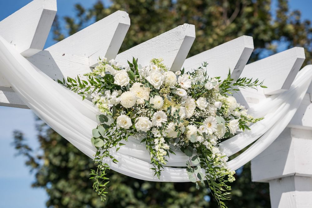 White floral arrangements adorn a wedding ceremony arch against a bright blue sky.