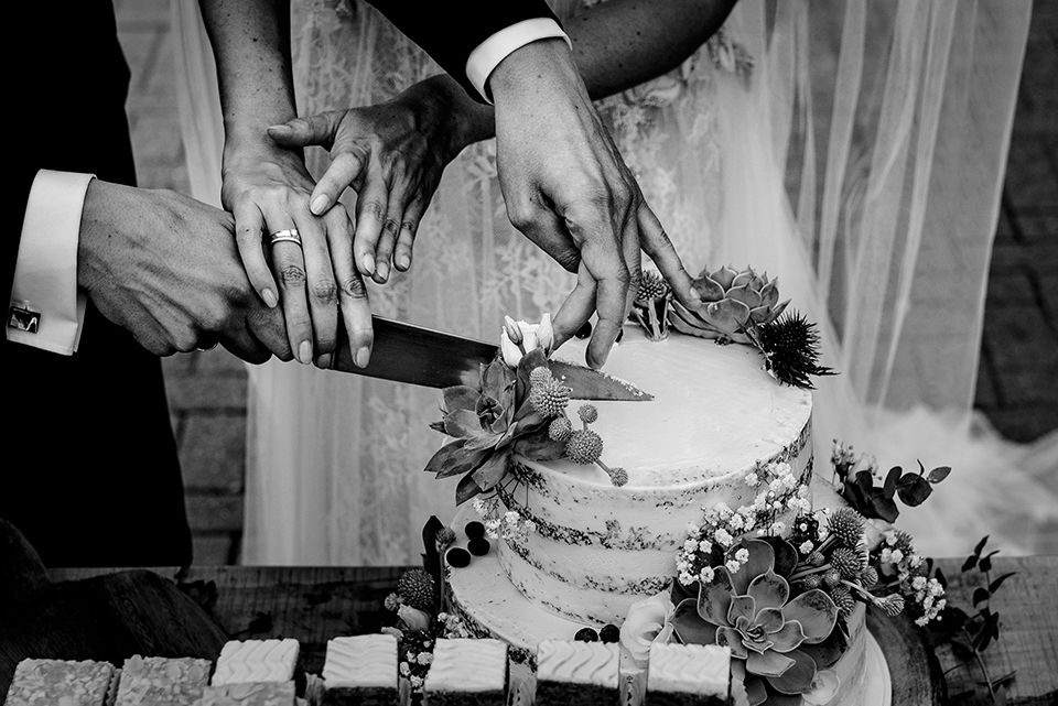 Black and white photo of hands cutting a wedding cake decorated with flowers.