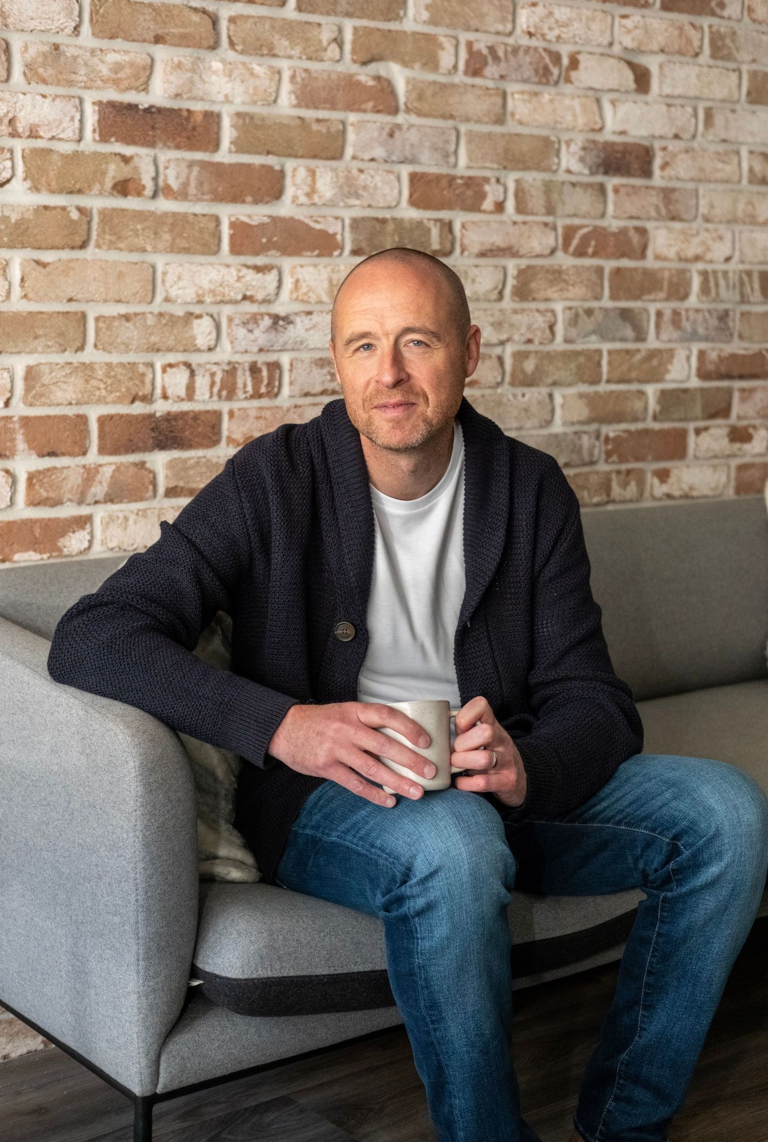A person in a black jacket and jeans sits casually on a gray couch against an exposed brick wall.