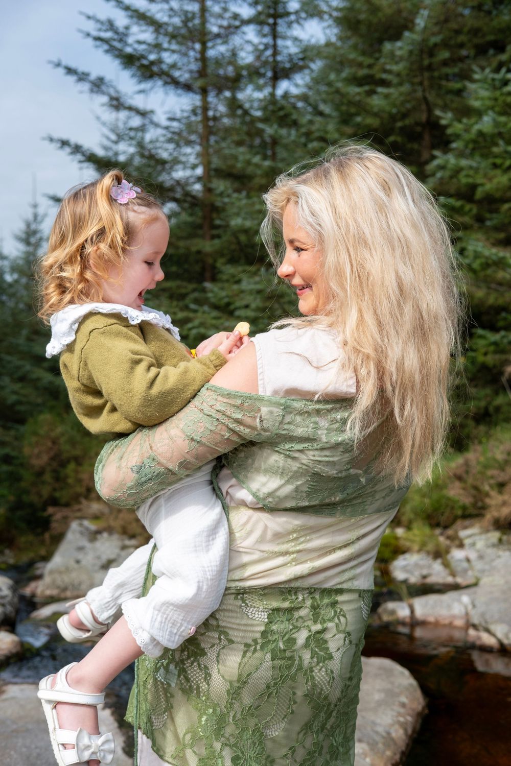 A parent and child share a joyful spinning moment together outdoors against a backdrop of evergreen trees.