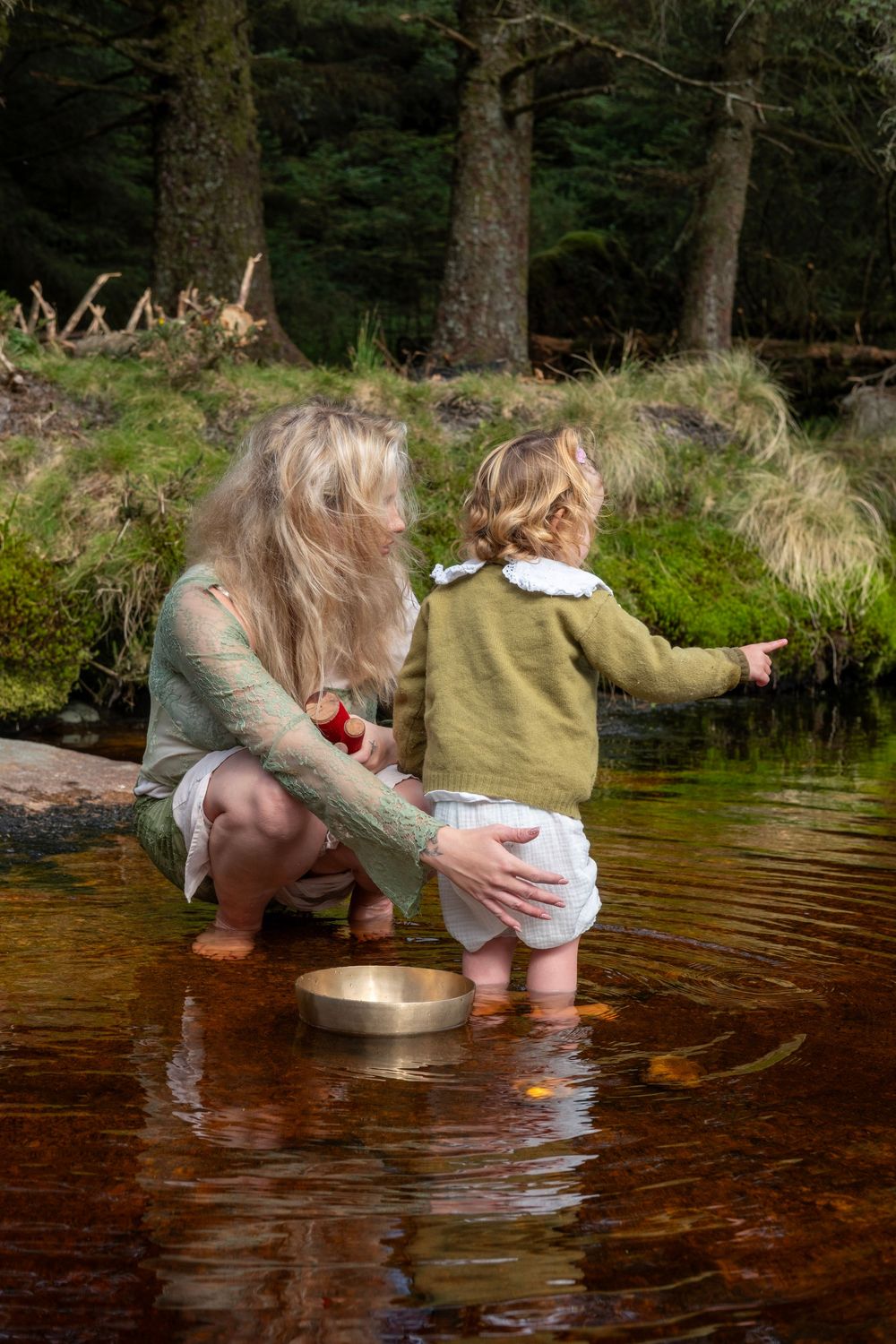 Two people in outdoor clothing kneel by a stream to collect water in a metal container from the brown-tinted water.