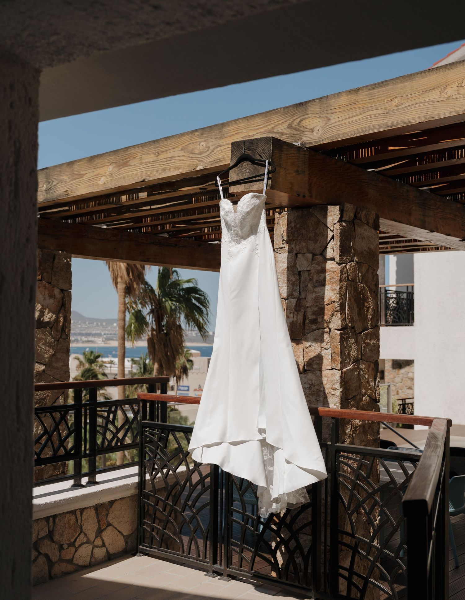 White wedding dress hanging on rustic stone balcony with palm trees and ocean view at tropical resort.
