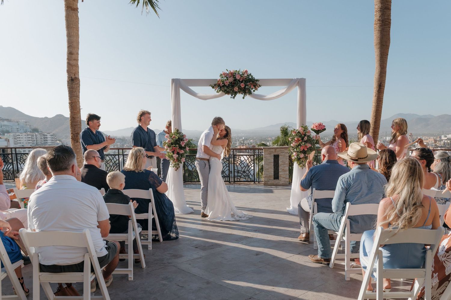 Beachfront wedding ceremony setup with white chairs and arch overlooking ocean under blue sky.