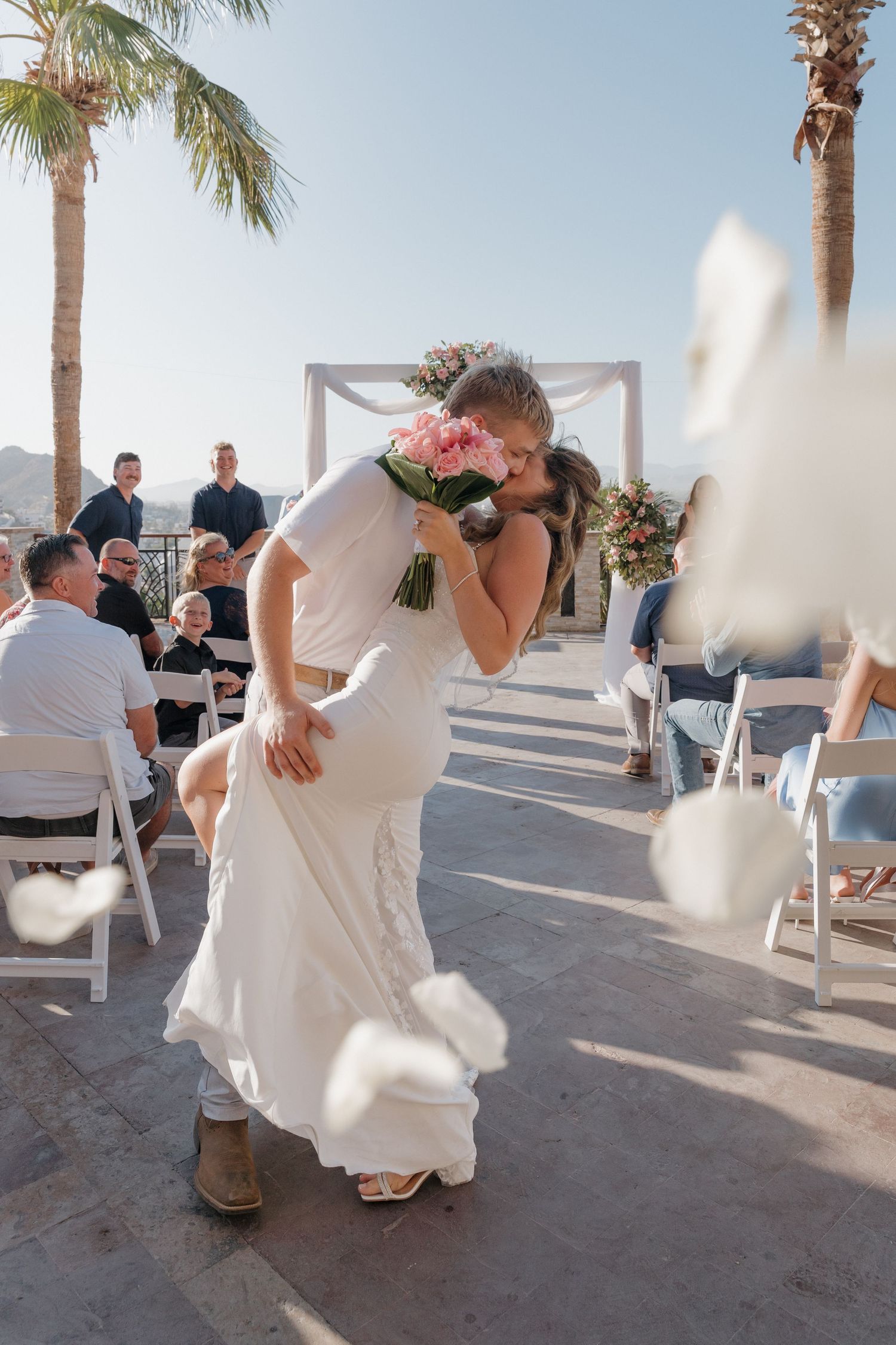 Wedding couple shares intimate moments during beach ceremony with palm trees and white chairs in the background.