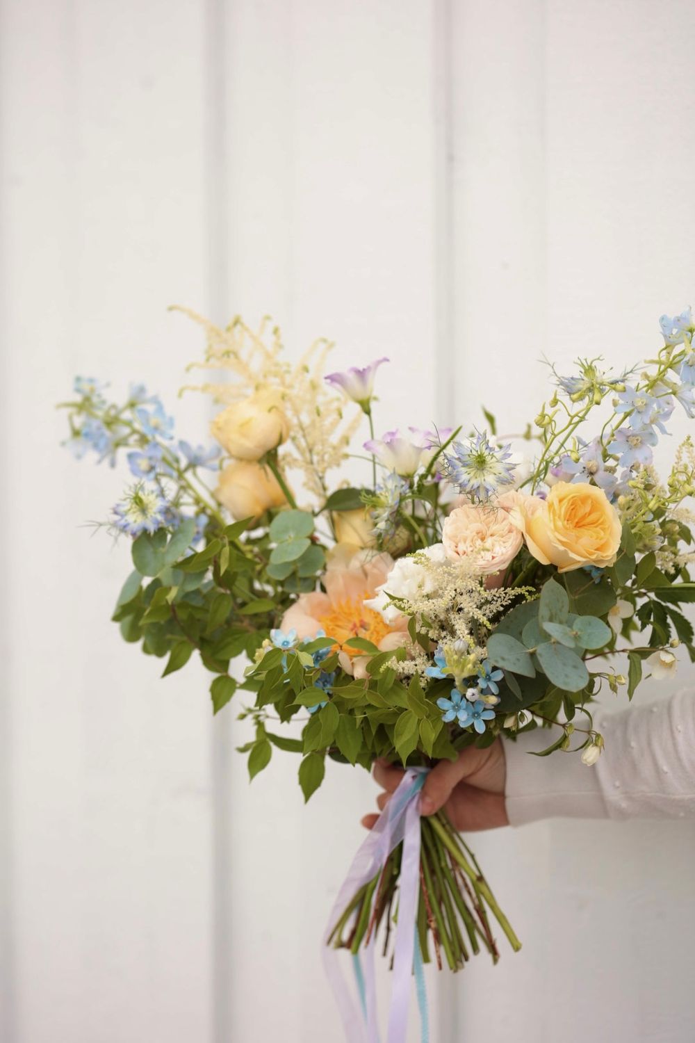 Hand holding a lush bridal bouquet with peach roses, blue wildflowers, and greenery tied with pastel ribbons.