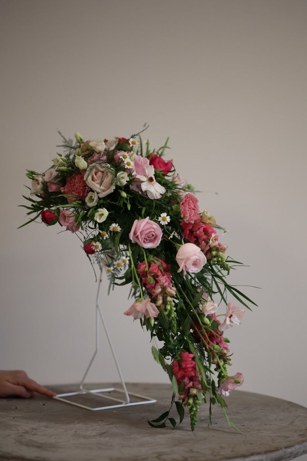 Pink roses and wildflowers cascading arrangement on white stand atop stone table against neutral wall.