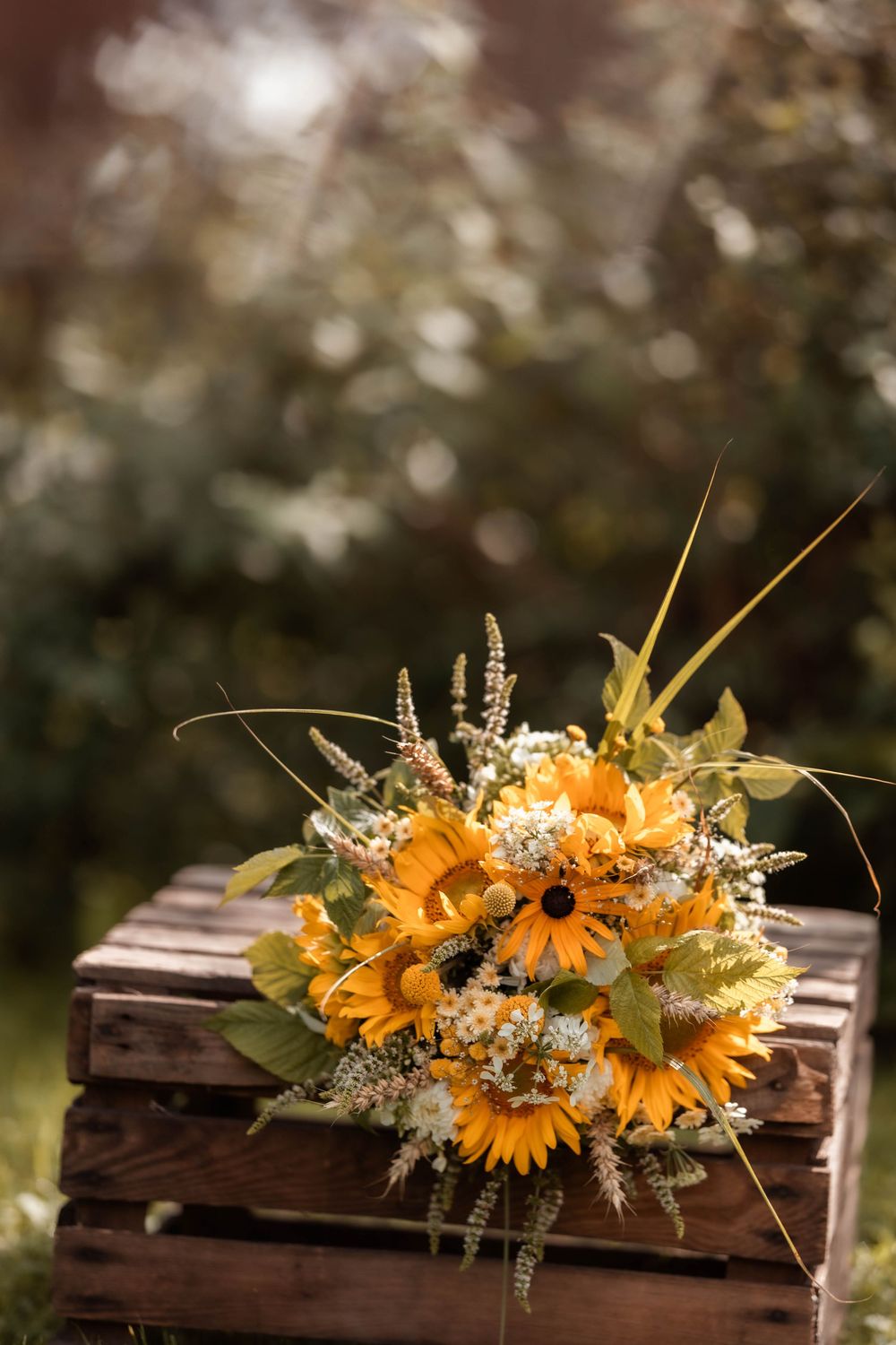 Rustic sunflower bouquet with wildflowers resting on a wooden crate outdoors with a soft green bokeh background.