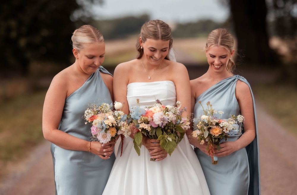 Bride in white gown with two bridesmaids in blue dresses holding floral bouquets on a scenic outdoor path.