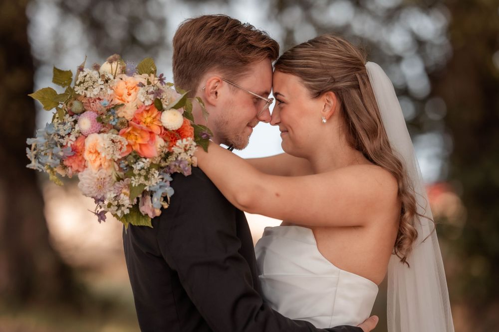 Bride and groom touching foreheads, smiling, with a peach and blue floral bouquet at an outdoor wedding.