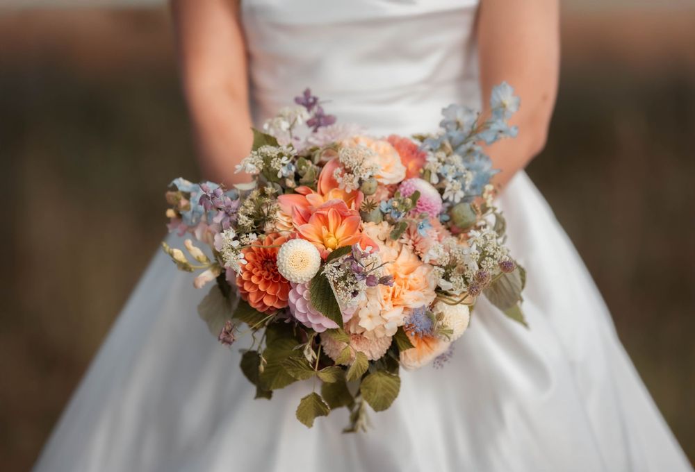 Bride in white gown holding colorful spring wedding bouquet with peach dahlias, lavender, and eucalyptus.