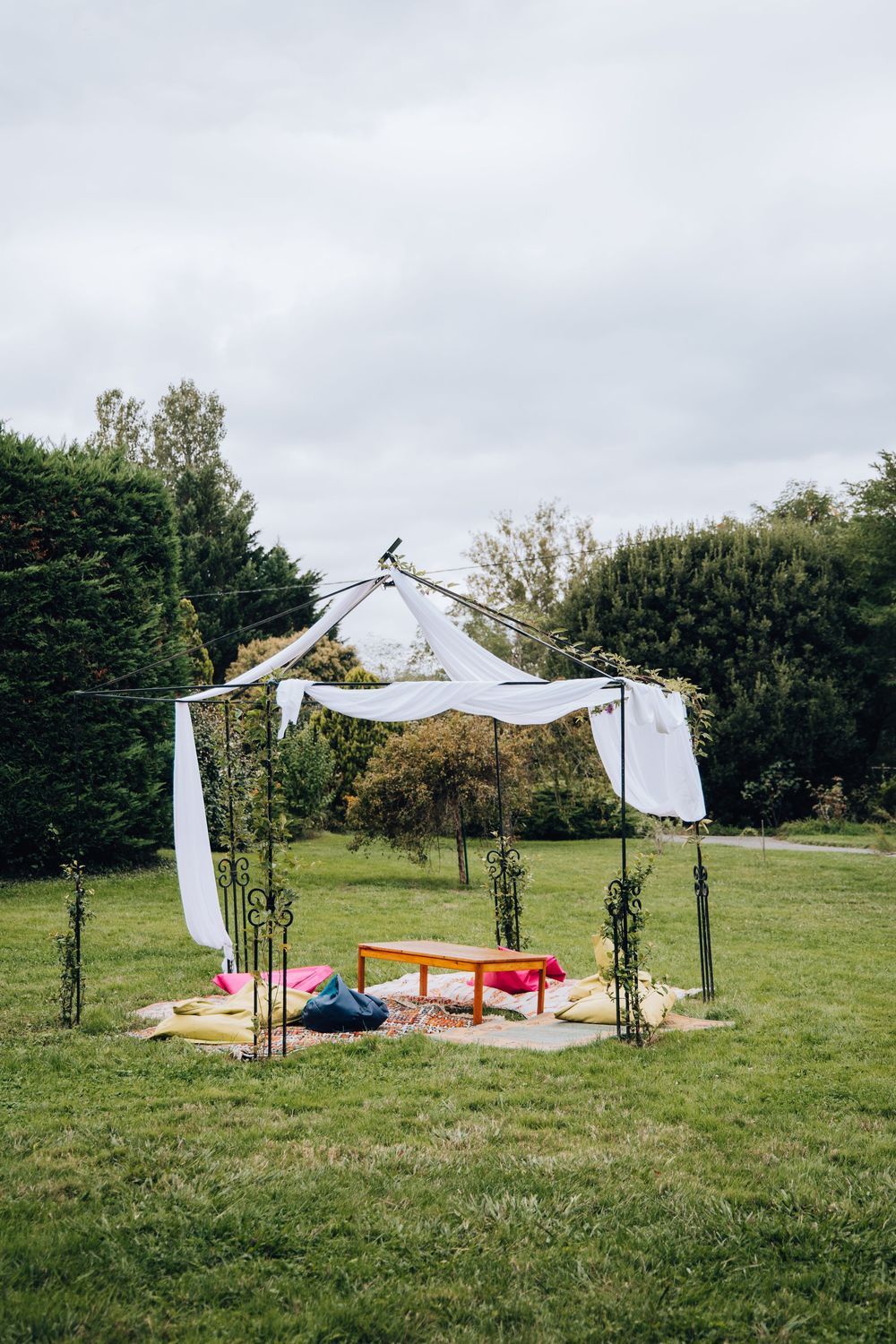 Outdoor wedding chuppah with white draped fabric canopy, colorful cushions and bench on green grass.