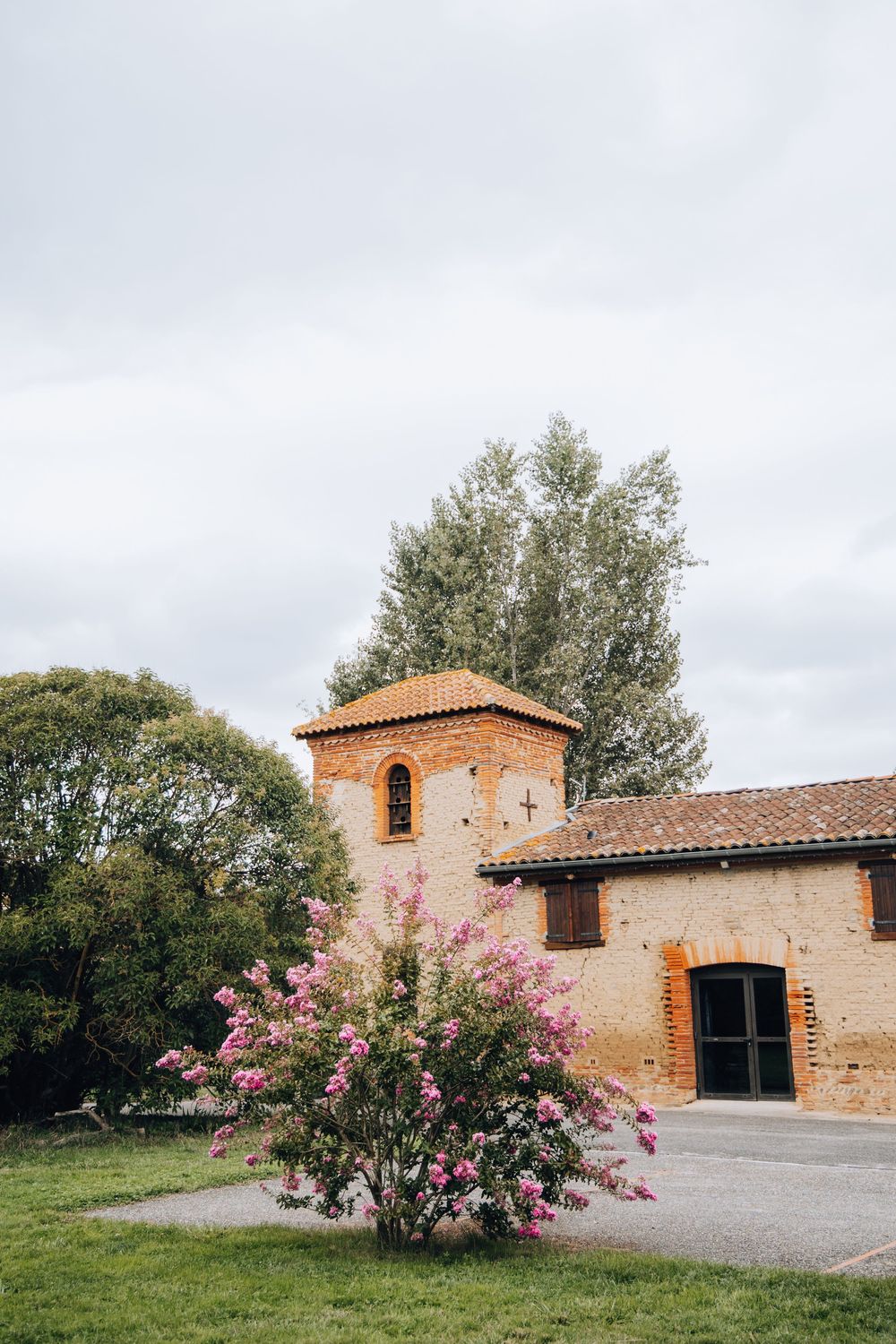 Rustic brick wedding venue with tower, terracotta roof, and blooming pink oleander bush in foreground.