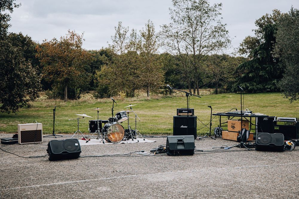 Outdoor live band stage setup with drum kit, amplifiers, and speakers on paved area overlooking green field.