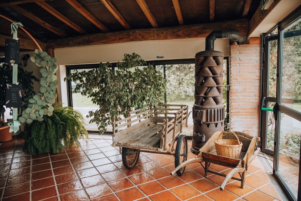 Cozy sunroom with terracotta tile floors, vintage wood stove, wicker chair, and lush green plants by large windows.