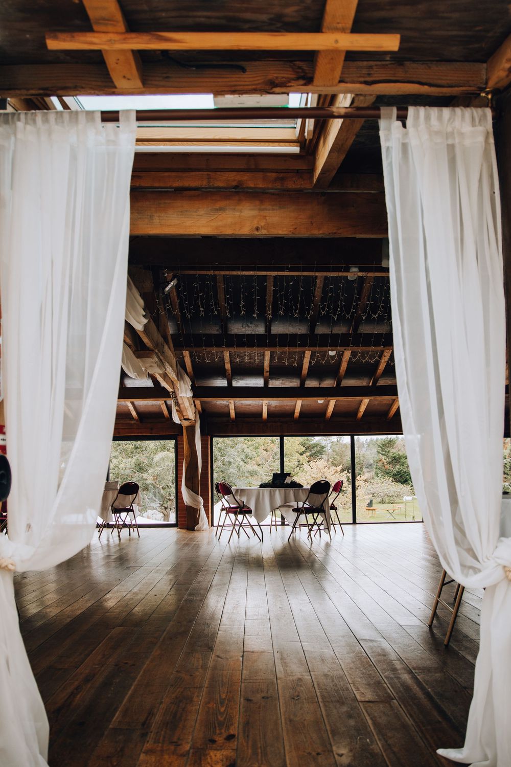 Rustic barn wedding venue interior with white sheer curtains, wooden beams, hardwood floors, and tables set for reception.