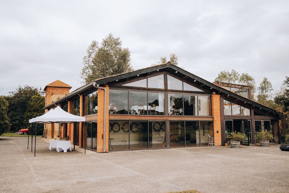 Modern barn-style wedding venue exterior with large glass windows, wooden accents, and white tent setup outside.